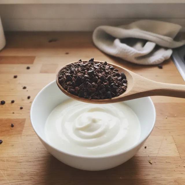 Cuillère en bois remplie d'éclats de grué de cacao au-dessus d'un bol de yaourt nature, sur un plan de travail de cuisine