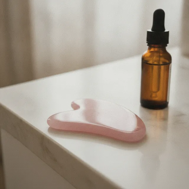 Gua sha en quartz rose posé sur un plan clair, à côté d'un flacon de sérum