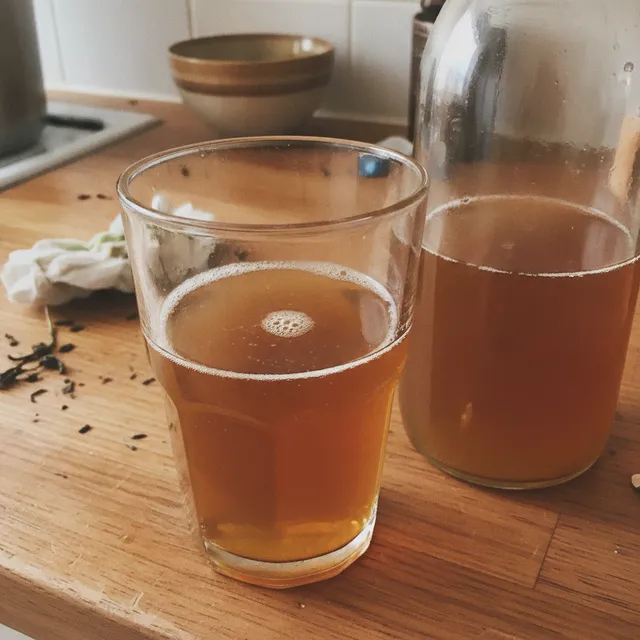 Un verre de kombucha ambre et petillant a cote d'une bouteille en verre sur un plan de travail en bois