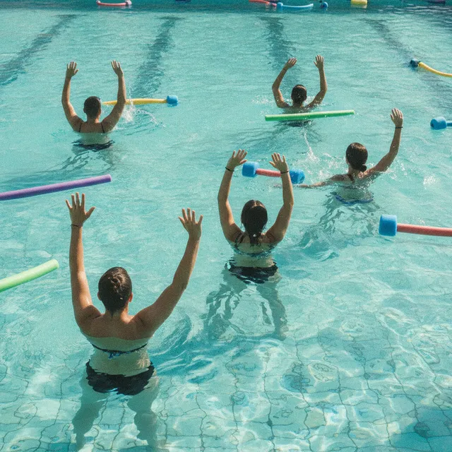 Cours d'aquagym dans une piscine, participants de dos les bras levés dans l'eau, frites en mousse flottantes