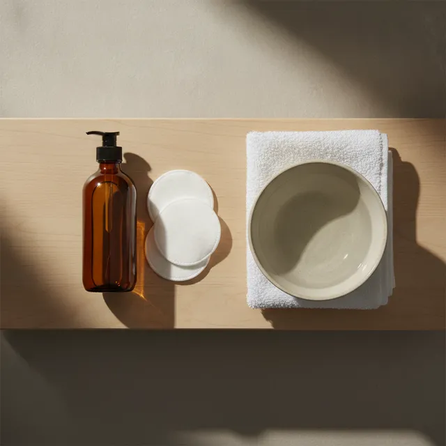 Flacon d'huile demaquillante, cotons lavables, serviette et bol d'eau sur une tablette de salle de bain en bois