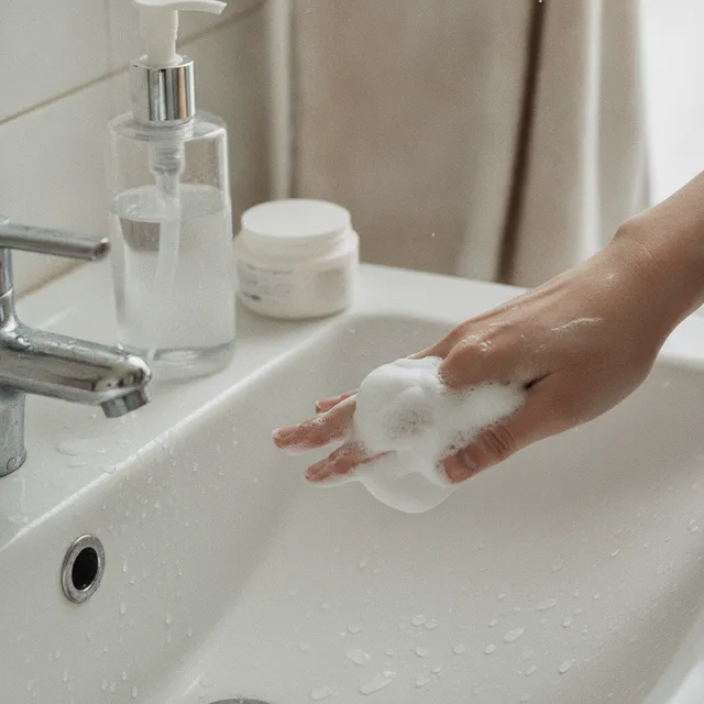 Une main fait mousser un nettoyant visage au-dessus d'un lavabo, flacon-pompe et pot posés sur le rebord