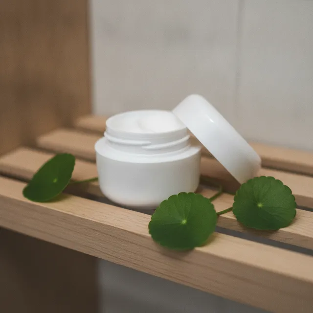Pot de crème cosmétique blanc entouré de feuilles rondes de centella asiatica, l'herbe du tigre