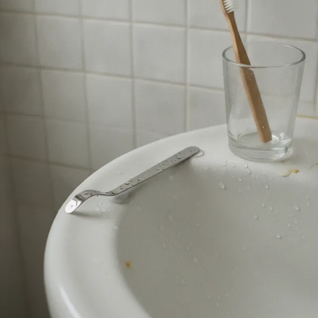 Un gratte-langue en inox posé sur le rebord d'un lavabo, à côté d'une brosse à dents