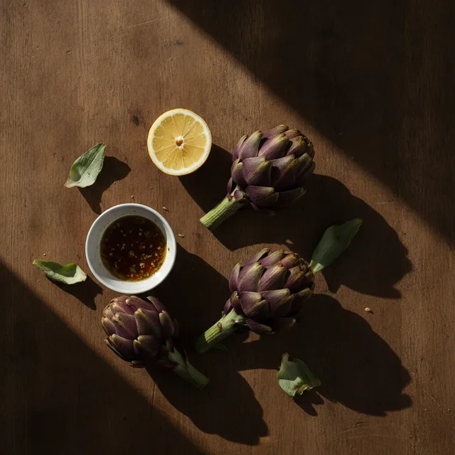 Trois artichauts violets posés sur une table en bois avec un demi-citron et un bol de vinaigrette, vue de dessus