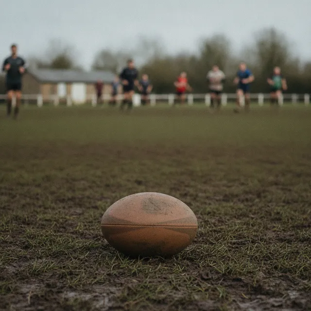 Ballon de rugby posé sur une pelouse boueuse, joueurs amateurs flous en arrière-plan
