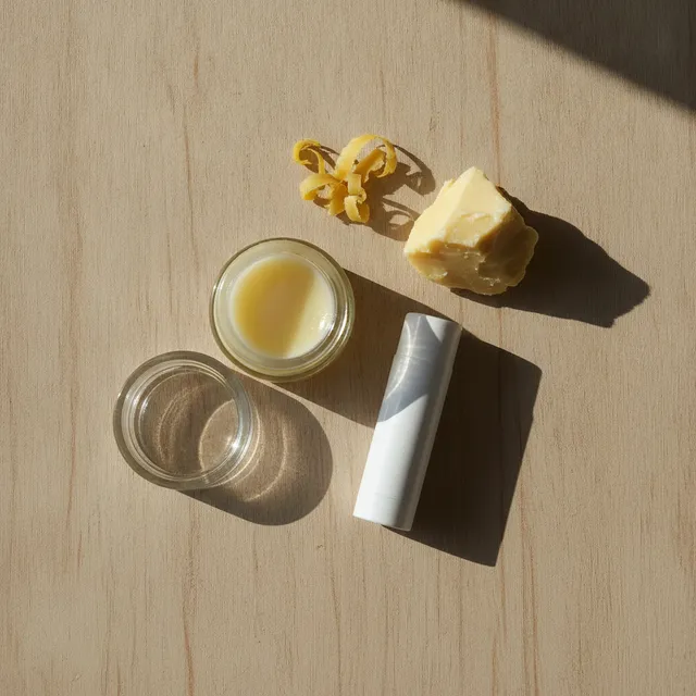 Un pot de baume à lèvres ouvert, un stick et un morceau de beurre de karité posés sur une table en bois.